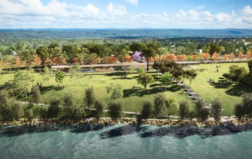 Regatta Park is a scenic riverside haven with a vast green lawn, scattered trees, and people sitting or walking. A paved path runs through the park precinct, lined with lush vegetation on both sides. In the background, a forest and distant hills rest under a partly cloudy blue sky.