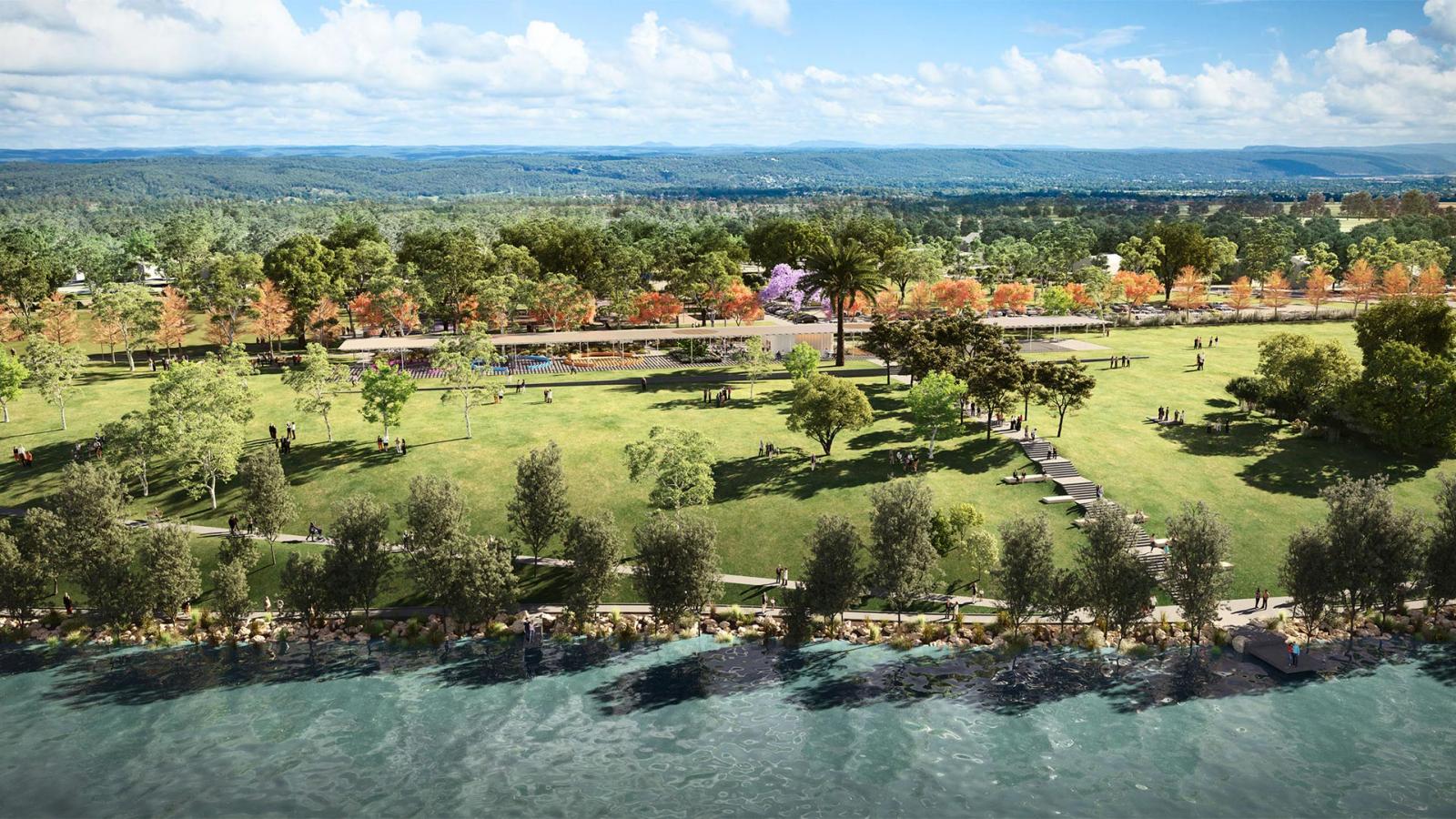 Regatta Park is a scenic riverside haven with a vast green lawn, scattered trees, and people sitting or walking. A paved path runs through the park precinct, lined with lush vegetation on both sides. In the background, a forest and distant hills rest under a partly cloudy blue sky.