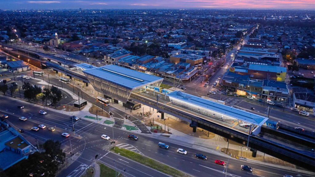 An aerial view of a modern elevated railway station in an urban setting, illuminated at dusk. The station has two platforms with trains aligned on both sides. Surrounding it are numerous buildings, streets, a few cars in motion, and even a nearby reservoir. The sky is tinged with a purple hue.