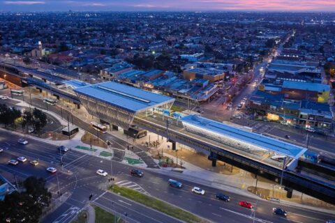 An aerial view of a modern elevated railway station in an urban setting, illuminated at dusk. The station has two platforms with trains aligned on both sides. Surrounding it are numerous buildings, streets, a few cars in motion, and even a nearby reservoir. The sky is tinged with a purple hue.