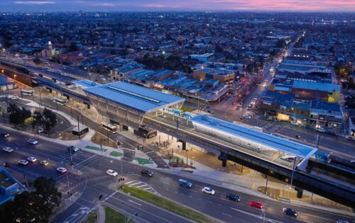 An aerial view of a modern elevated railway station in an urban setting, illuminated at dusk. The station has two platforms with trains aligned on both sides. Surrounding it are numerous buildings, streets, a few cars in motion, and even a nearby reservoir. The sky is tinged with a purple hue.