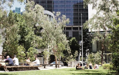 A group of people enjoy a sunny day in an urban park surrounded by trees and modern buildings at Quay Place. Some sit at benches and tables, talking and relaxing, while others sit on the grass, creating a lively and social atmosphere amidst the greenery by the riverside cityscape.