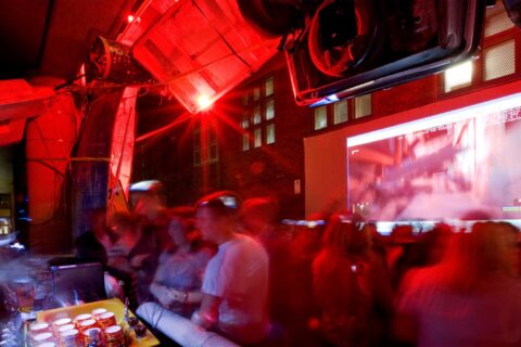 A dimly lit, red-tinted room filled with a large crowd of people, possibly at a party or event. The Seven Metre Bar counter on the left boasts various drinks, and a blurred projection screen on the wall displays an indiscernible image. The atmosphere appears lively.