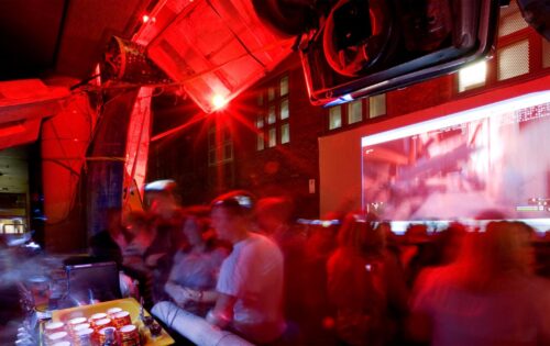 A dimly lit, red-tinted room filled with a large crowd of people, possibly at a party or event. The Seven Metre Bar counter on the left boasts various drinks, and a blurred projection screen on the wall displays an indiscernible image. The atmosphere appears lively.