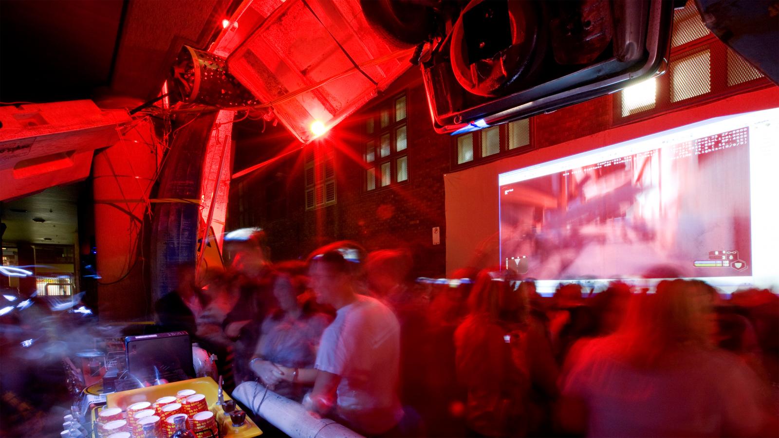 A dimly lit, red-tinted room filled with a large crowd of people, possibly at a party or event. The Seven Metre Bar counter on the left boasts various drinks, and a blurred projection screen on the wall displays an indiscernible image. The atmosphere appears lively.