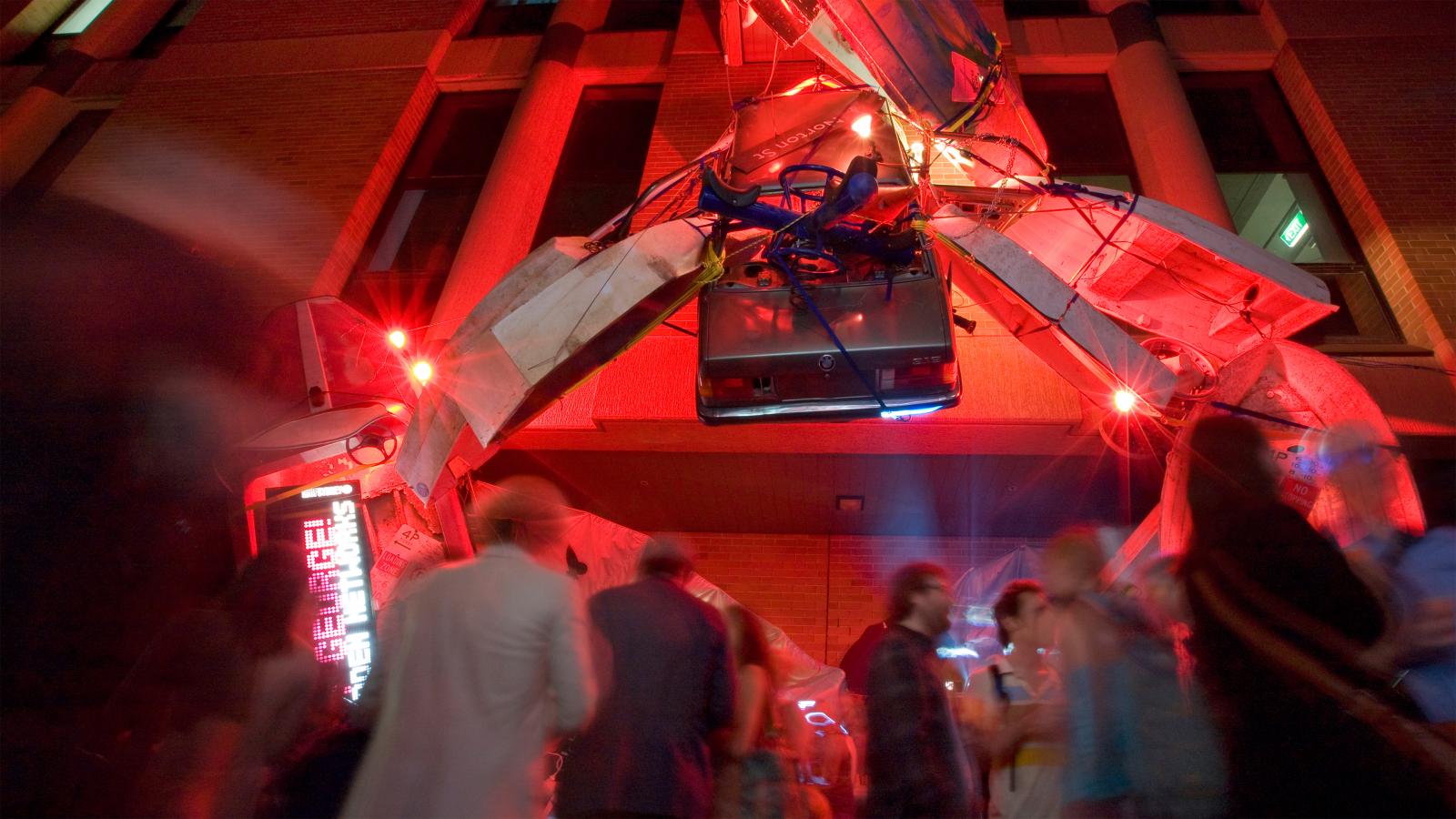 A large, surreal art installation with red lighting is suspended above a crowd of blurry, moving people. The structure, which includes what looks like electronic waste, looms over the vibrant Seven Metre Bar. The scene is vivid and lively, suggesting a night event or festival.