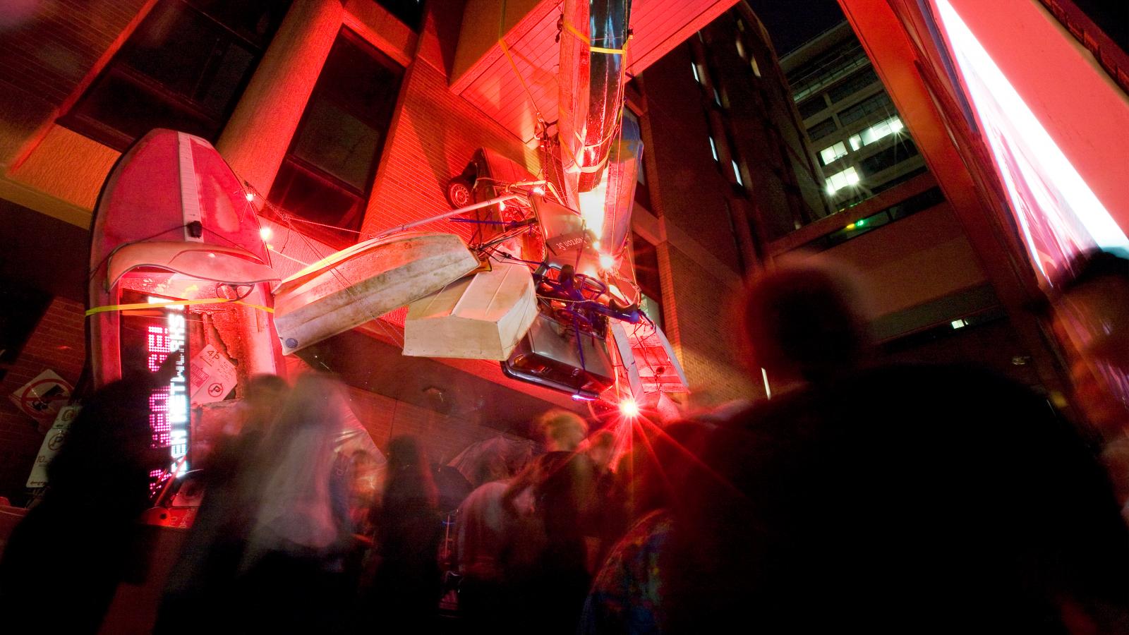 A vibrant street scene at night features an abstract installation of various objects, including boats and planks, lit by intense red lighting near the Seven Metre Bar in an urban setting. Blurred silhouettes of people move around, creating a dynamic atmosphere.