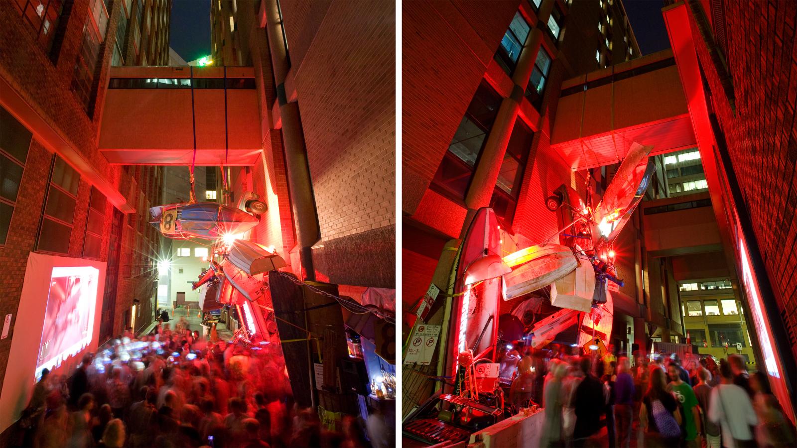 A crowded alley lit in red features an art installation with airplanes and vehicles hanging between buildings. On the left, people gather near a large screen by the Seven Metre Bar; on the right, they observe the suspended sculptures. The scene is lively and illuminated by city lights.