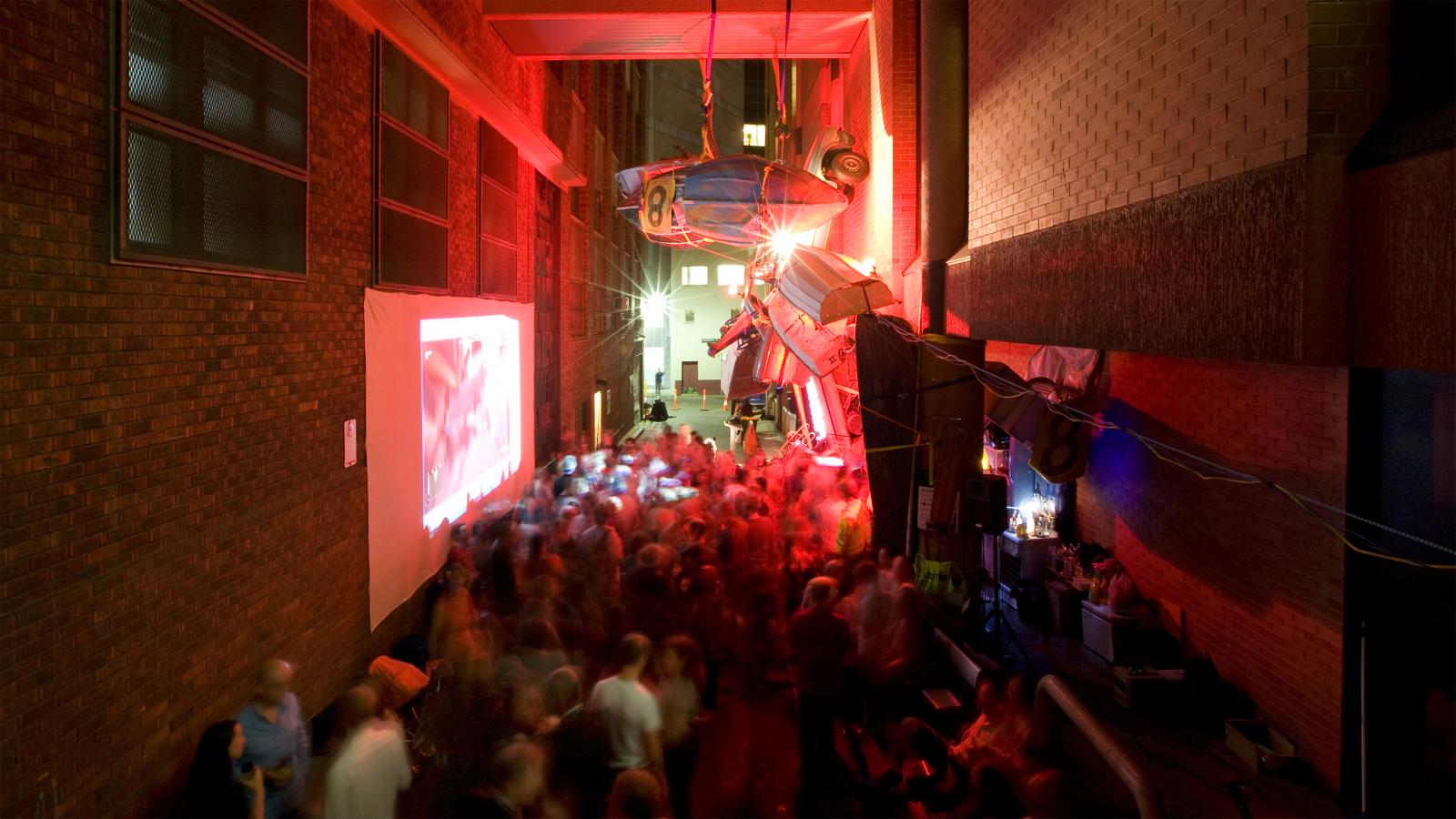 A lively night scene in an alleyway filled with a crowd of people. Bright, colorful lights illuminate the area, highlighting a large projection screen on one side and vehicles suspended mid-air above the festive Seven Metre Bar. The atmosphere is energetic and buzzing with excitement.