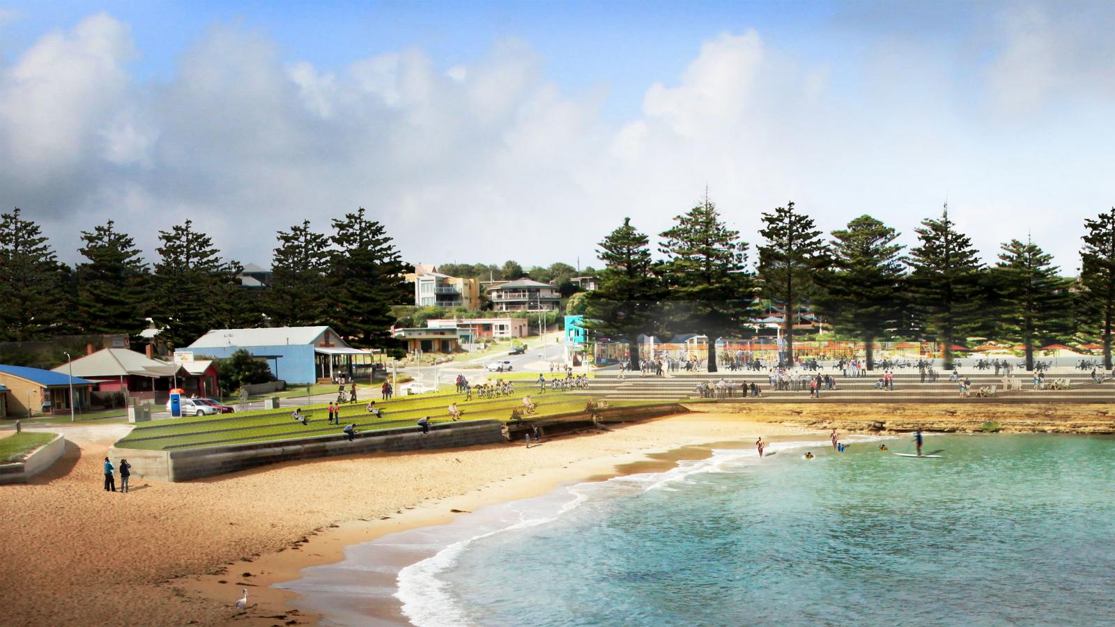 A scenic coastal view featuring the picturesque Shipwreck Coast with a sandy beach where people enjoy the water and shoreline. Green grassy areas in the foreground have visitors sitting and walking. The background showcases buildings, large trees, and more people strolling near the shoreline.