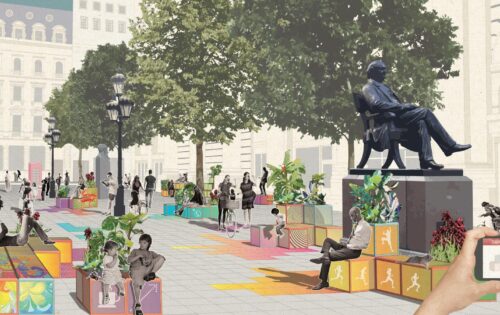 A vibrant urban plaza with people sitting and walking among colorful, decorated blocks. Trees line the path, providing shade. A large statue of a seated figure is prominent. Someone stands on a smart carpet, holding a smartphone in the foreground, capturing the lively scene.