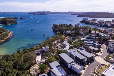 Aerial view of Spring Cove Manly, featuring a coastal residential area with modern houses and flat roofs. Below, clear blue waters with boats sailing and wooded areas are visible. In the distance, a cityscape with tall buildings under a slightly cloudy sky completes the serene scene.
