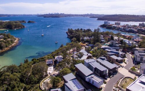 Aerial view of Spring Cove Manly, featuring a coastal residential area with modern houses and flat roofs. Below, clear blue waters with boats sailing and wooded areas are visible. In the distance, a cityscape with tall buildings under a slightly cloudy sky completes the serene scene.