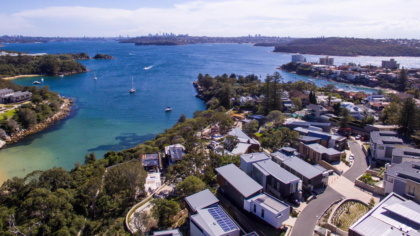 Aerial view of Spring Cove Manly, featuring a coastal residential area with modern houses and flat roofs. Below, clear blue waters with boats sailing and wooded areas are visible. In the distance, a cityscape with tall buildings under a slightly cloudy sky completes the serene scene.