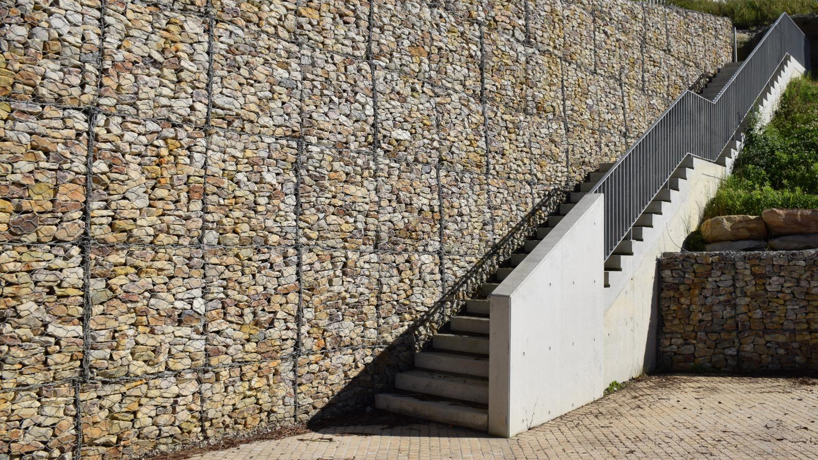 A concrete staircase with gray railings is built against a large stone retaining wall made of rocks enclosed in wire cages. The steps lead up to a higher level with greenery visible on the right side, reminiscent of Manly’s Spring Cove. The ground is paved with tiles.