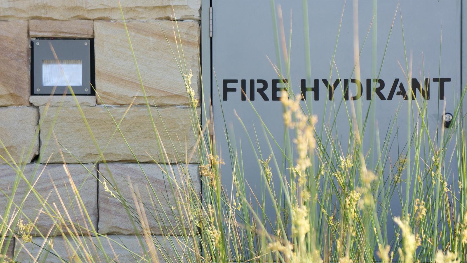 A gray fire hydrant door is positioned next to a tan stone wall along Spring Cove. Tall grasses grow in the foreground, partially obscuring the door and wall. A small framed sign is attached to the wall just above some of the grasses, evoking a sense of abiding nature amidst urban manly surroundings.