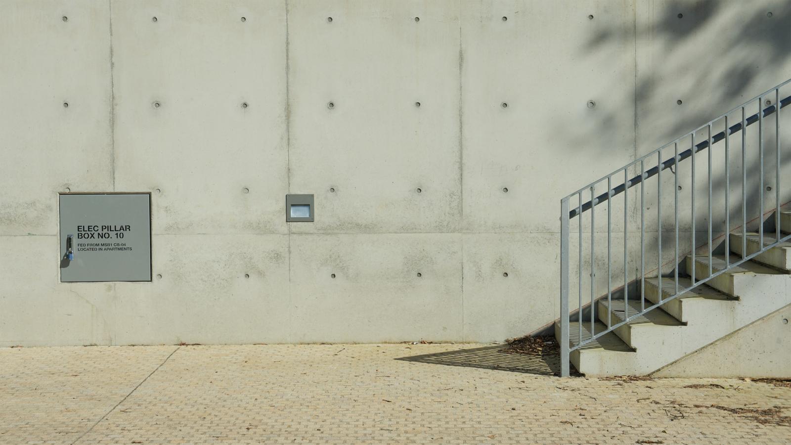 A minimalist, manly outdoor scene featuring a concrete wall with a small door labeled "ELEC PILAR (BLOCK NO. 10)" and an electrical warning. Beside it, a small light fixture and a metal staircase casting a shadow on the ground. The area, reminiscent of Spring Cove, is paved with light-colored tiles.
