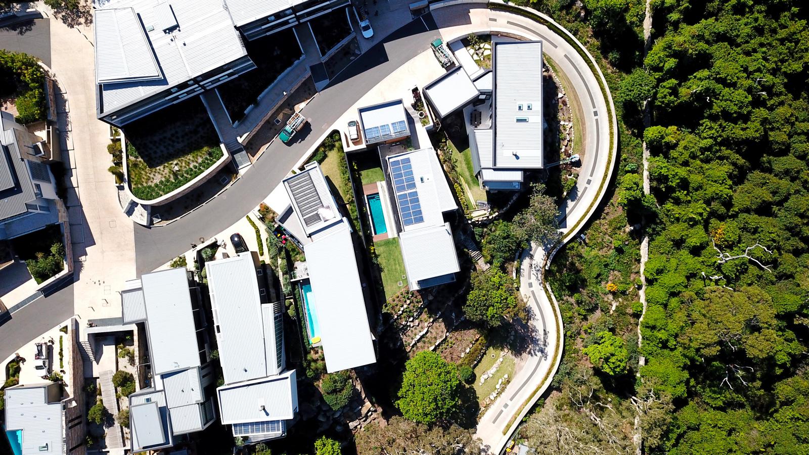 Aerial view of the modern residential neighborhood of Spring Cove, featuring several white-roofed houses. Some homes boast swimming pools. A winding road separates the neighborhood from a densely forested area, while neat, parallel rows of houses enhance the tranquility of Manly's scenic beauty.