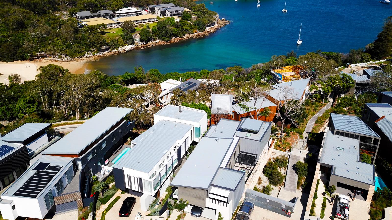 Aerial view of a coastal neighborhood in Spring Cove, featuring modern homes with flat roofs and large windows. The area, near Manly, is surrounded by lush greenery and overlooks a serene, blue waterfront with several boats anchored. There's a sandy beach area adjacent to the water.