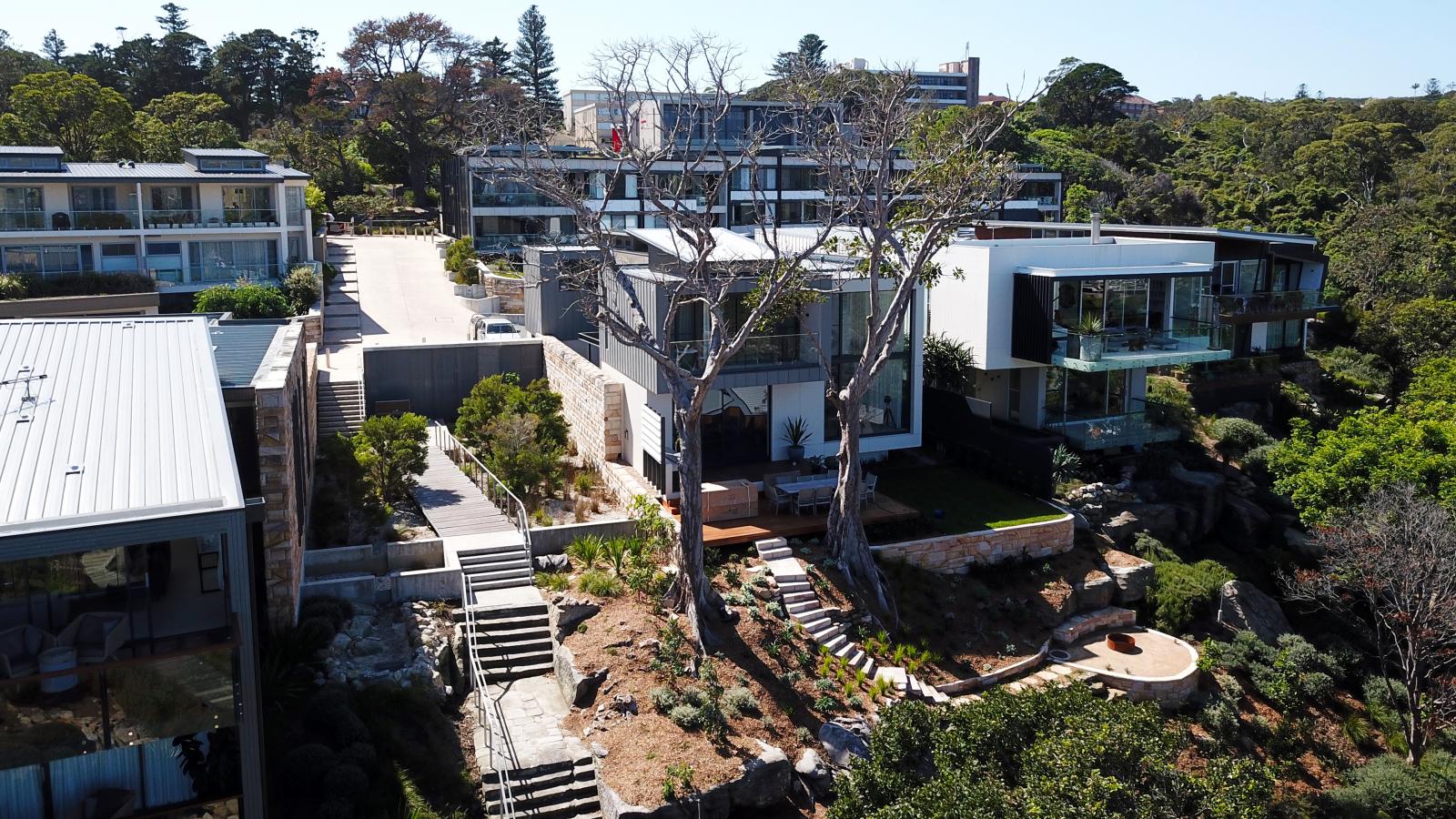 Aerial view of modern multi-story homes with large glass windows and balconies in Manly's Spring Cove, surrounded by lush greenery. A set of stairs leads up to the houses, and the area features various landscaped plants and trees.