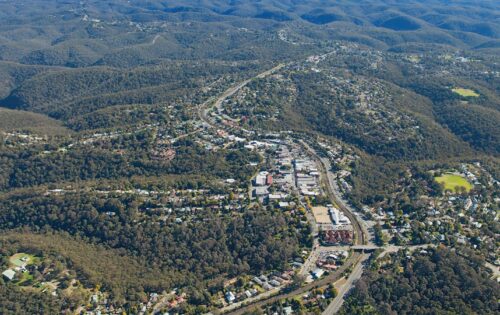 Aerial view of Springwood Town Centre nestled in a lush, hilly landscape surrounded by dense forest. The town features a masterplan with a network of roads, residential and commercial buildings, and a prominent highway cutting through the scene. The area is encompassed by rolling, tree-covered hills.