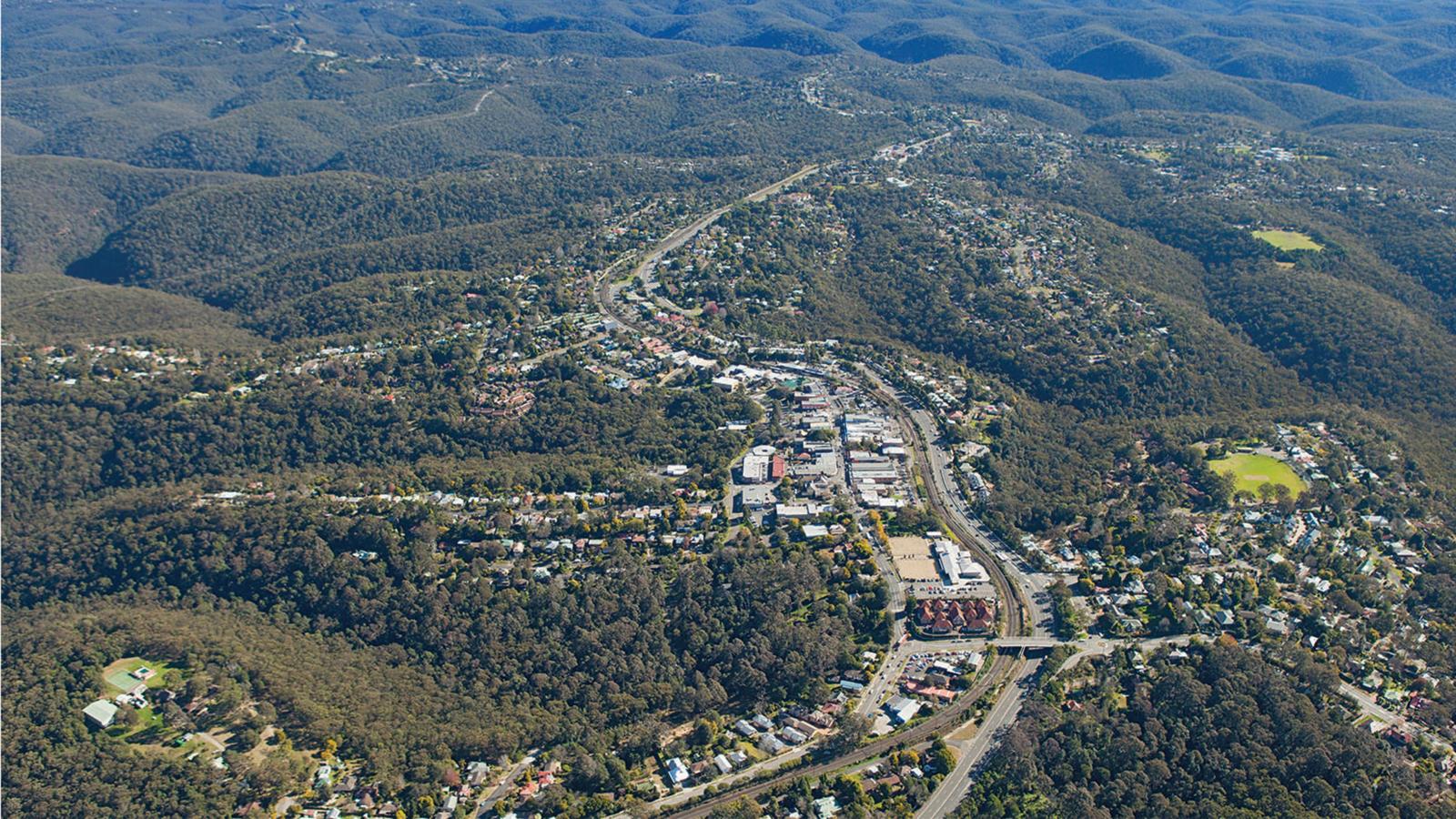 Aerial view of Springwood Town Centre nestled in a lush, hilly landscape surrounded by dense forest. The town features a masterplan with a network of roads, residential and commercial buildings, and a prominent highway cutting through the scene. The area is encompassed by rolling, tree-covered hills.