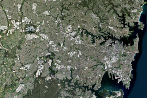 A satellite image of a densely populated urban area featuring a mix of grey urban structures and green spaces. Water bodies are visible along the right edge, suggesting a coastal location like Sydney. Roads and buildings form intricate grid patterns, indicating a large cityscape.