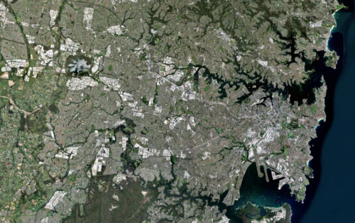 A satellite image of a densely populated urban area featuring a mix of grey urban structures and green spaces. Water bodies are visible along the right edge, suggesting a coastal location like Sydney. Roads and buildings form intricate grid patterns, indicating a large cityscape.