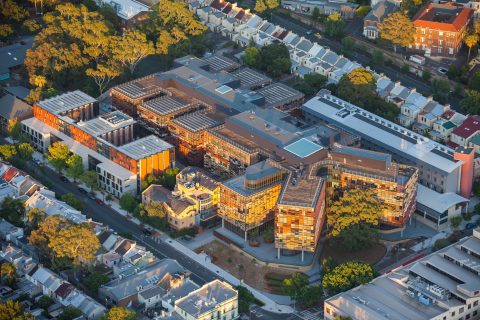 Aerial view of the University of Sydney's expansive, modern building complex, surrounded by lush green trees and a residential neighborhood with uniform row houses. The business school within has interconnected structures with glass facades and rooftop solar panels. Streets frame the perimeter.