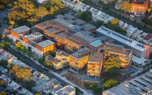 Aerial view of the University of Sydney's expansive, modern building complex, surrounded by lush green trees and a residential neighborhood with uniform row houses. The business school within has interconnected structures with glass facades and rooftop solar panels. Streets frame the perimeter.