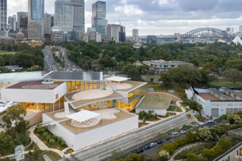 Aerial view of a modern, architecturally unique building with multiple levels and terraces surrounded by greenery. City skyline, roads, Sydney Harbour Bridge, and Sydney Opera House visible in the background under a cloudy sky. Discover more about us through this stunning urban landscape.