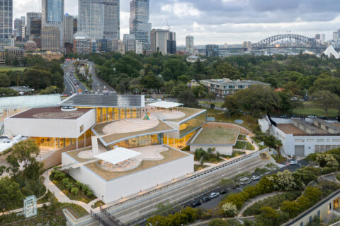 Aerial view of the Sydney Modern Project, an expansion of the Art Gallery of New South Wales, showcasing contemporary architectural design with rooftop terraces and green spaces. The iconic Sydney Harbour Bridge and Sydney Opera House enhance the breathtaking backdrop.