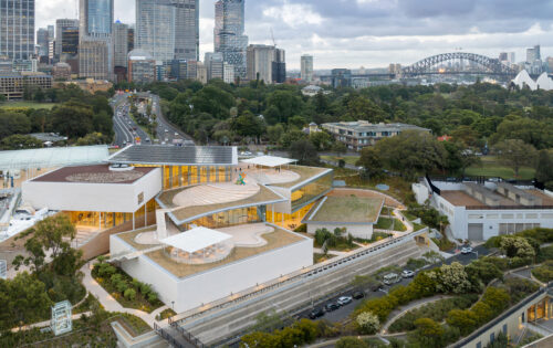 Aerial view of the Sydney Modern Project, an expansion of the Art Gallery of New South Wales, showcasing contemporary architectural design with rooftop terraces and green spaces. The iconic Sydney Harbour Bridge and Sydney Opera House enhance the breathtaking backdrop.