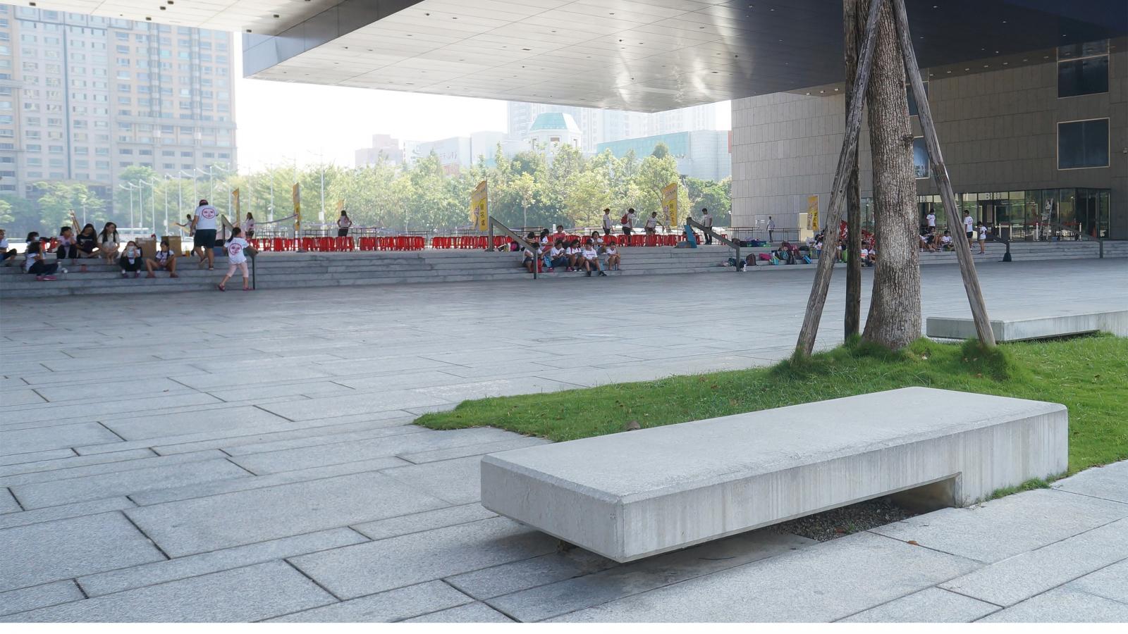 A modern urban plaza in Taichung features a large, angular building with overhanging architecture. Sparse groups of people sit or walk around the open area, where trees and greenery are interspersed among the paved sections. Red chairs set up in rows suggest preparation for a government event.