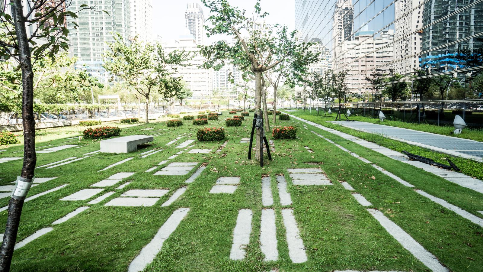 A modern park in Taichung features neatly arranged green lawns and pathways made of rectangular stone slabs. Trees and shrubs are interspersed throughout, while high-rise glass buildings reflect the greenery. The scene is bright and sunny, perfect for enjoying this beautifully planned Stage 1 development.