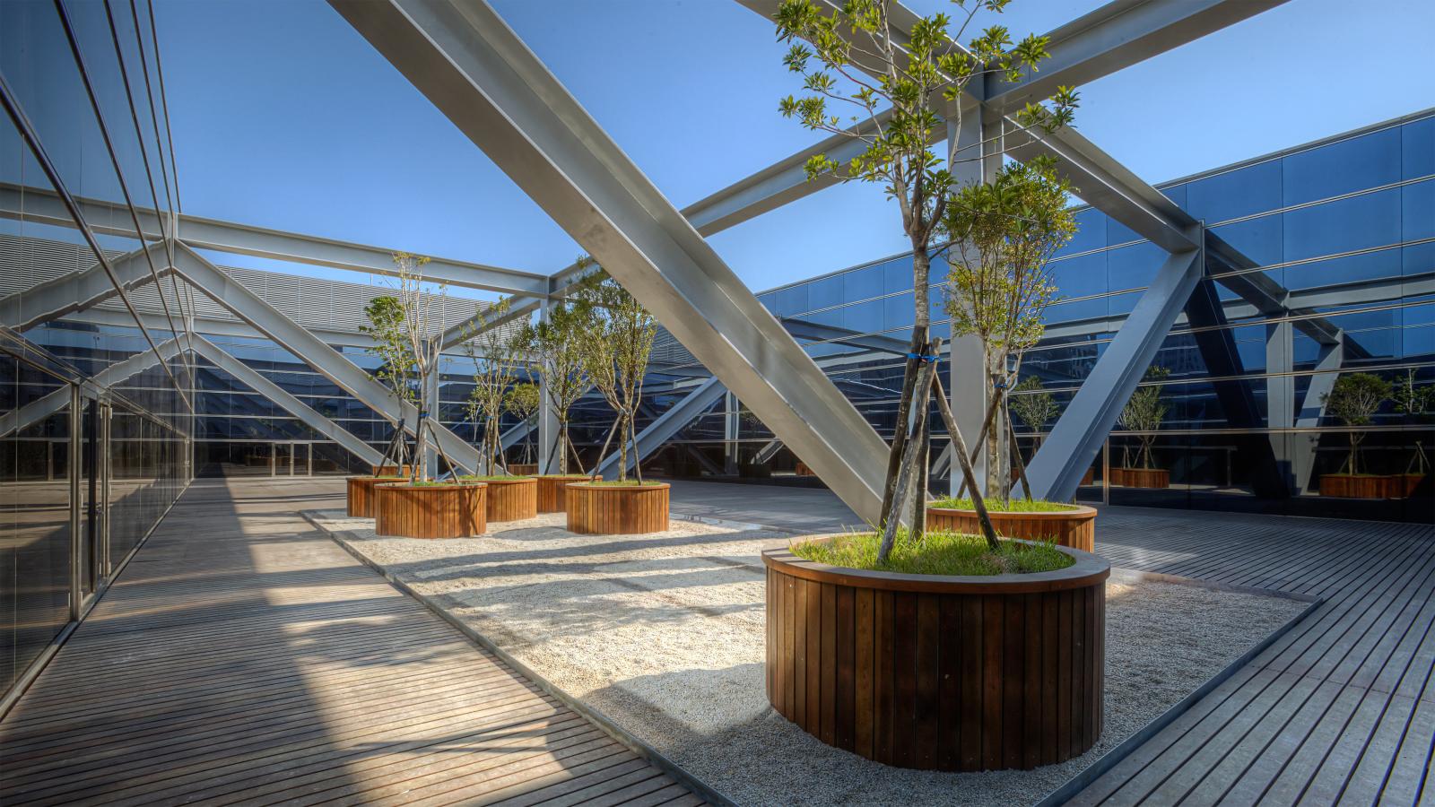 A modern courtyard with wooden planters holding young trees, set between sleek glass buildings in Taichung. The courtyard features wooden decking and white gravel. Large metal beams crisscross overhead, creating geometric patterns. This space marks Stage 1 of the government project under a clear, blue sky.