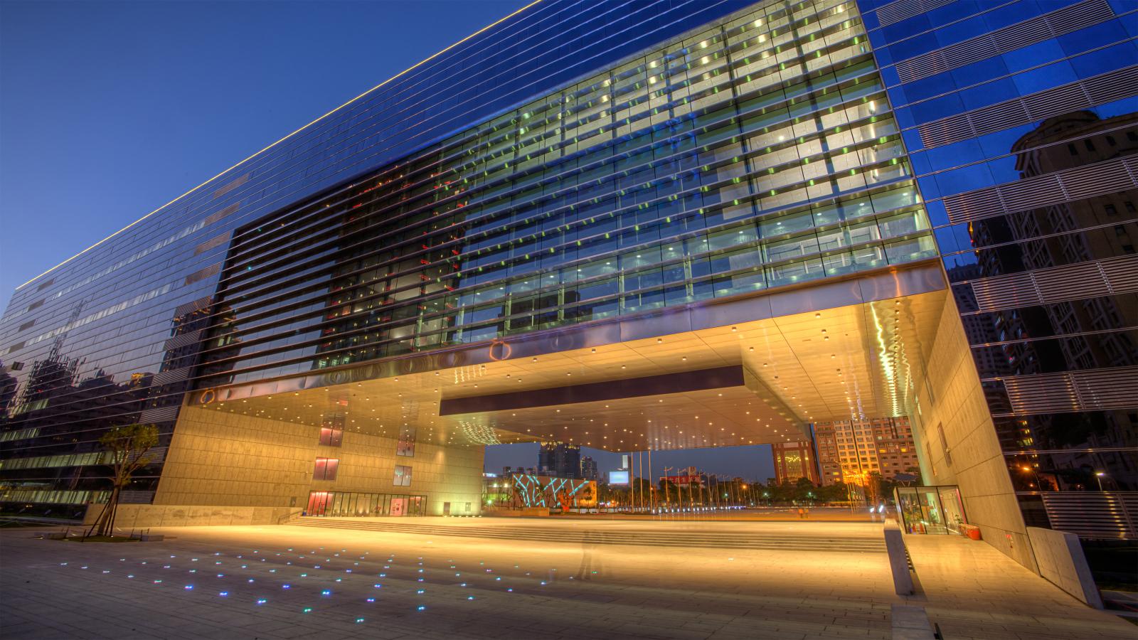 A modern glass and metal building with a large open archway in the middle stands as Stage 1 of Taichung’s new government complex. The building reflects the evening sky, and colorful lights are embedded in the ground. Inside the archway, there are illuminated panels and reflections of surrounding structures.