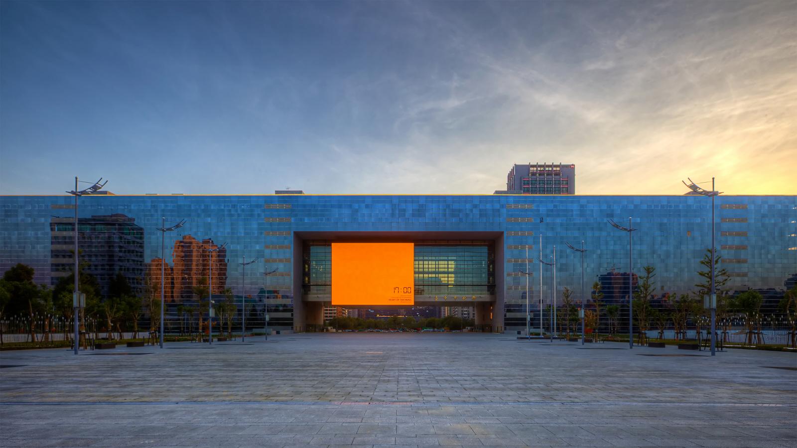 A large, modern building with a reflective glass facade is centered in the image, located in the heart of Taichung. In the middle of the building, a large, square orange screen displays text. Multiple streetlights and a plaza with tiled flooring lead up to the entrance. The sky above is partly cloudy.