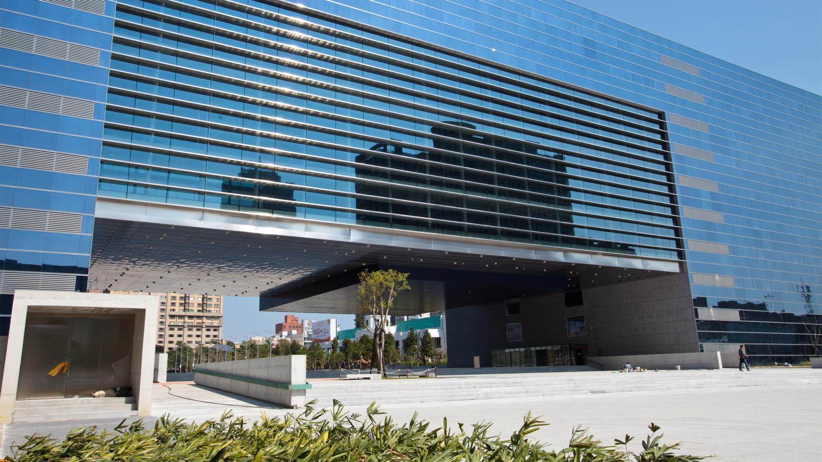 A modern glass building in Taichung with a large overhanging structure reflects nearby buildings on its windows. The foreground features a plaza with scattered greenery and a pathway leading to the entrance, reminiscent of government architecture in its Stage 1 development. The sky is clear and blue.