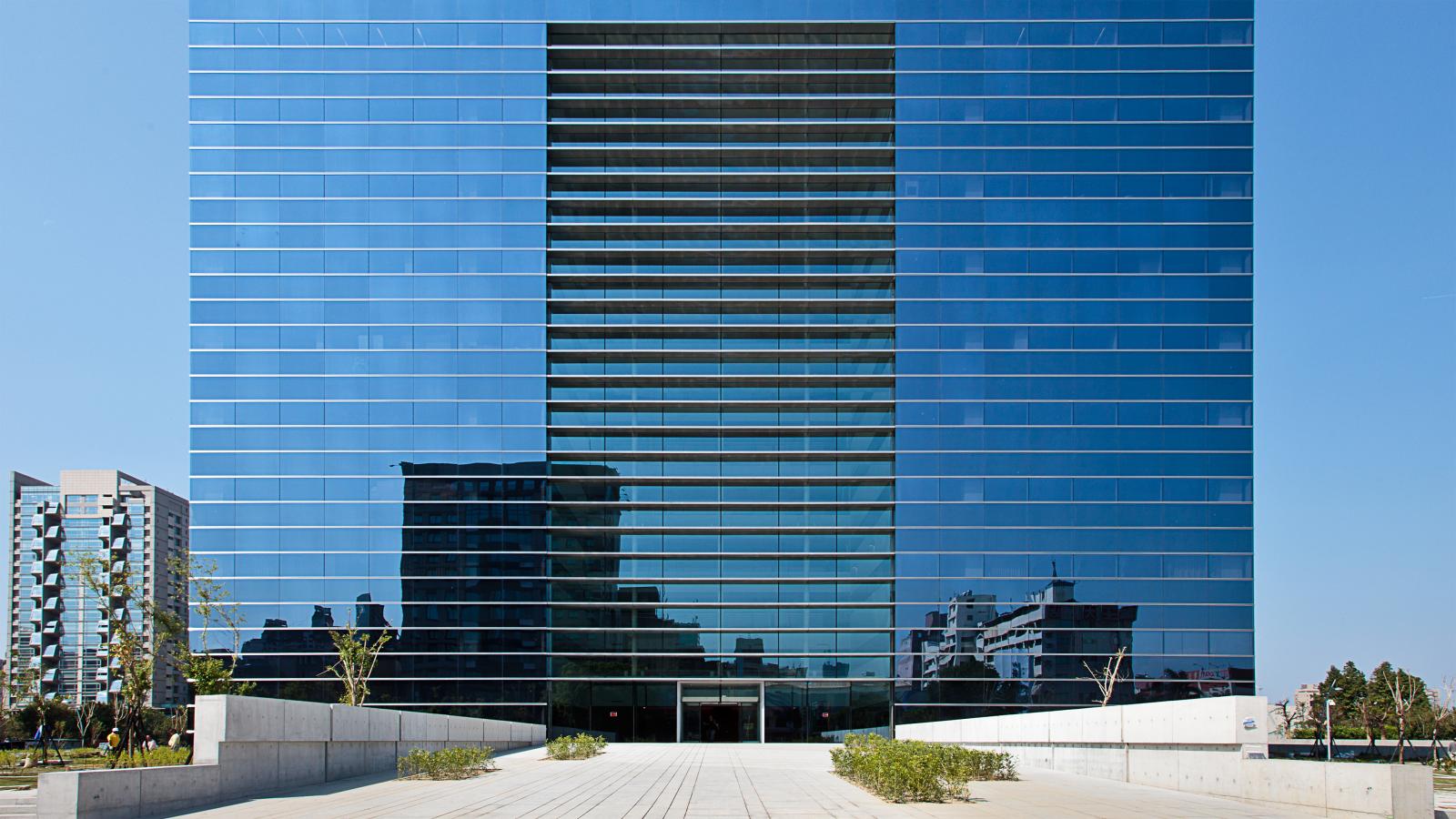 A modern glass office building with a tall, reflective facade stands at the center of Taichung's Stage 1 development. The entrance area features concrete pathways and sparse landscaping. The background shows additional buildings and a clear, blue sky.