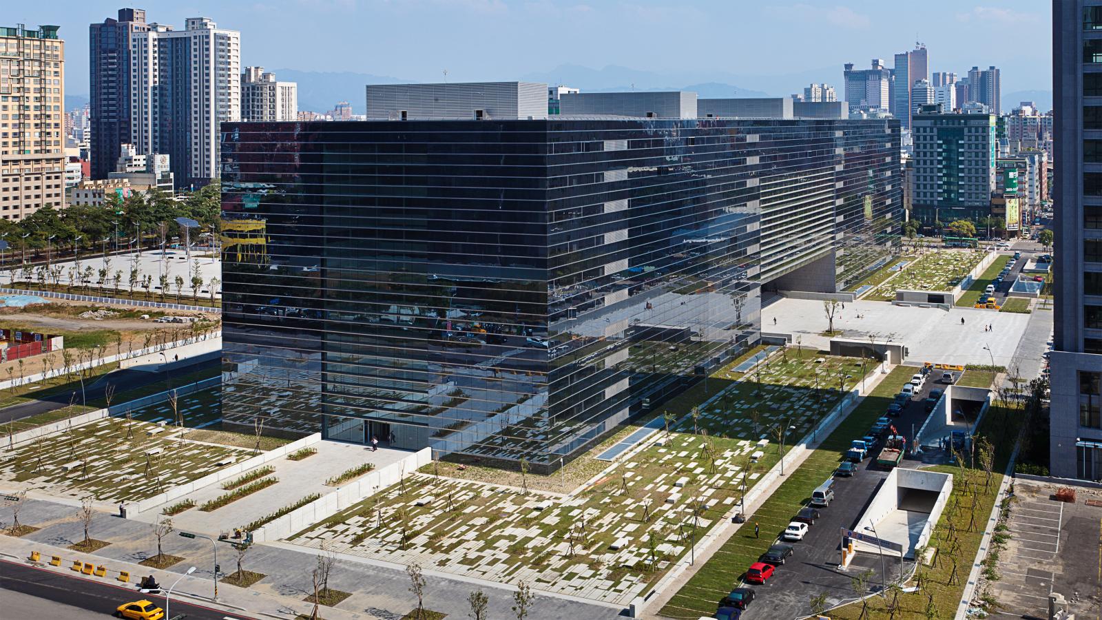 A large, modern, multi-story building with a sleek, black glass exterior sits in an urban area of Taichung. The surrounding landscape features neatly arranged patches of grass and pathways. Other buildings and a street with cars are visible in the background, indicating it's just Stage 1 of development.