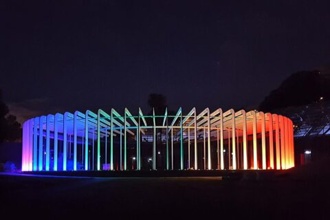 A circular structure at the Royal Botanic Gardens, known as the Calyx, is illuminated at night with vibrant, multi-colored lights transitioning from blue and purple on the left, to green and yellow in the center, and to orange and red on the right, creating a striking visual effect.