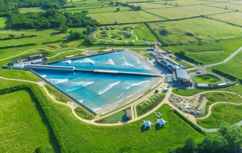 Aerial view of The Wave London, a large wave pool facility surrounded by green fields and trees. The site features a man-made wave generator creating waves, a viewing area, several small buildings, pathways, two tent structures, and a parking lot.