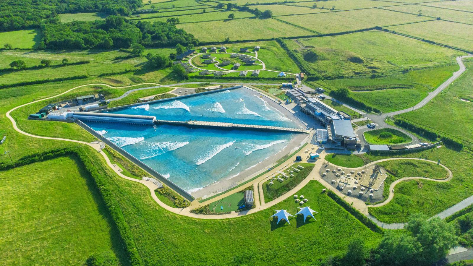 Aerial view of The Wave London, a large wave pool facility surrounded by green fields and trees. The site features a man-made wave generator creating waves, a viewing area, several small buildings, pathways, two tent structures, and a parking lot.