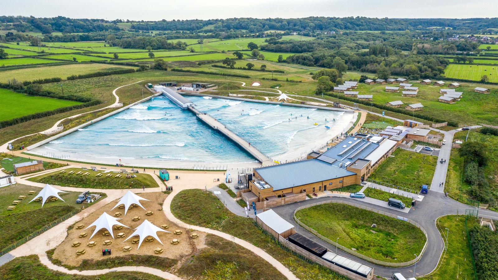 Aerial view of The Wave, a large artificial surfing lagoon in a rural area near London, surrounded by green fields and trees. The lagoon features consistent waves and is enclosed by a white barrier. Nearby are modern buildings, pathways, and several white canopy structures for shade.