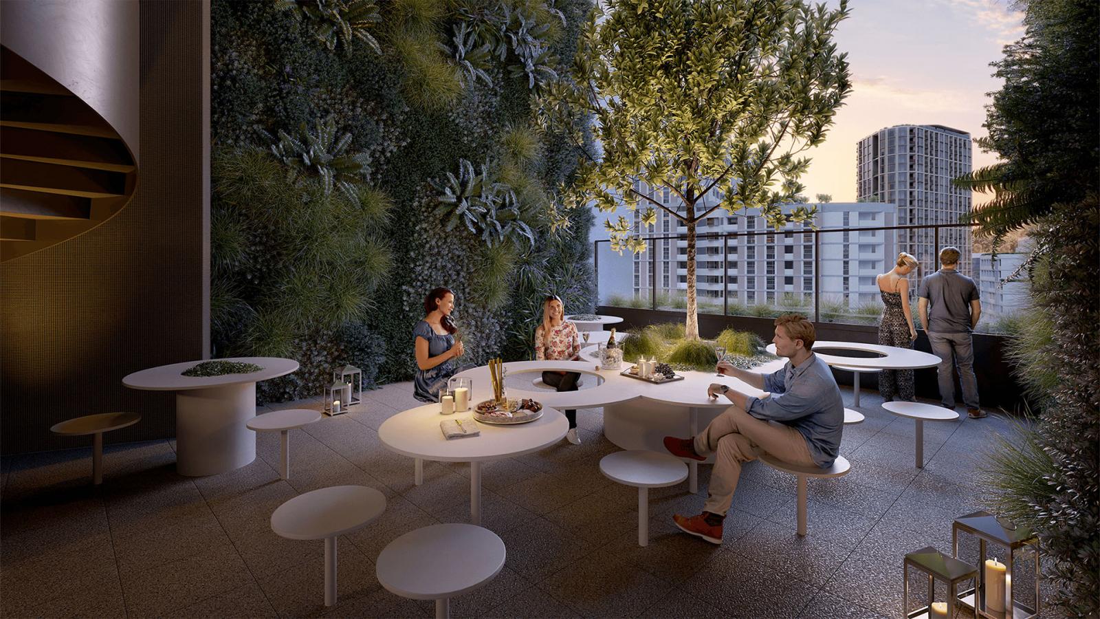 People are sitting and standing around modern, circular tables on a rooftop terrace with greenery on the walls and a tree in the center. One person is using a laptop, enjoying the evening ambiance of the Ivanhoe Estate treehouse-like setting with the cityscape visible in the background.