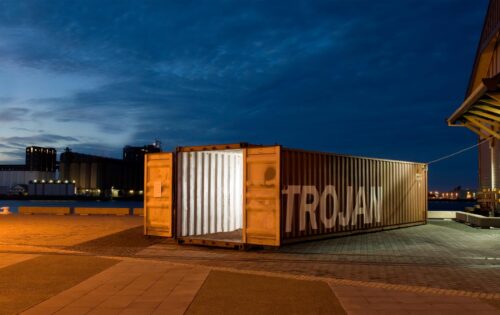 A large shipping container with the word "TROJAN" written on its side is illuminated from within, sitting on a paved area near a waterfront at dusk. The background shows industrial buildings and a calm water body under a deep blue sky, evoking thoughts of heading back to the city.