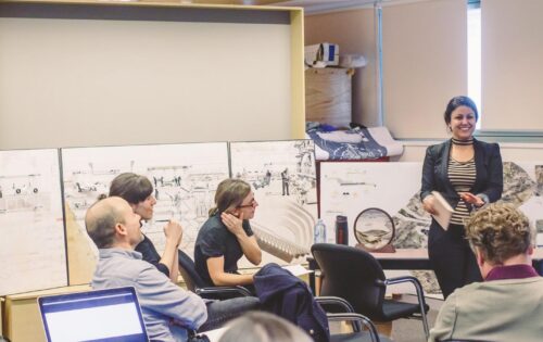 A group of people are gathered in a meeting room at the UTS Design Studio. One person stands and presents with a smile, while the others sit and listen attentively. Behind them are display boards with various MArch diagrams and sketches. The room has large windows and blinds.