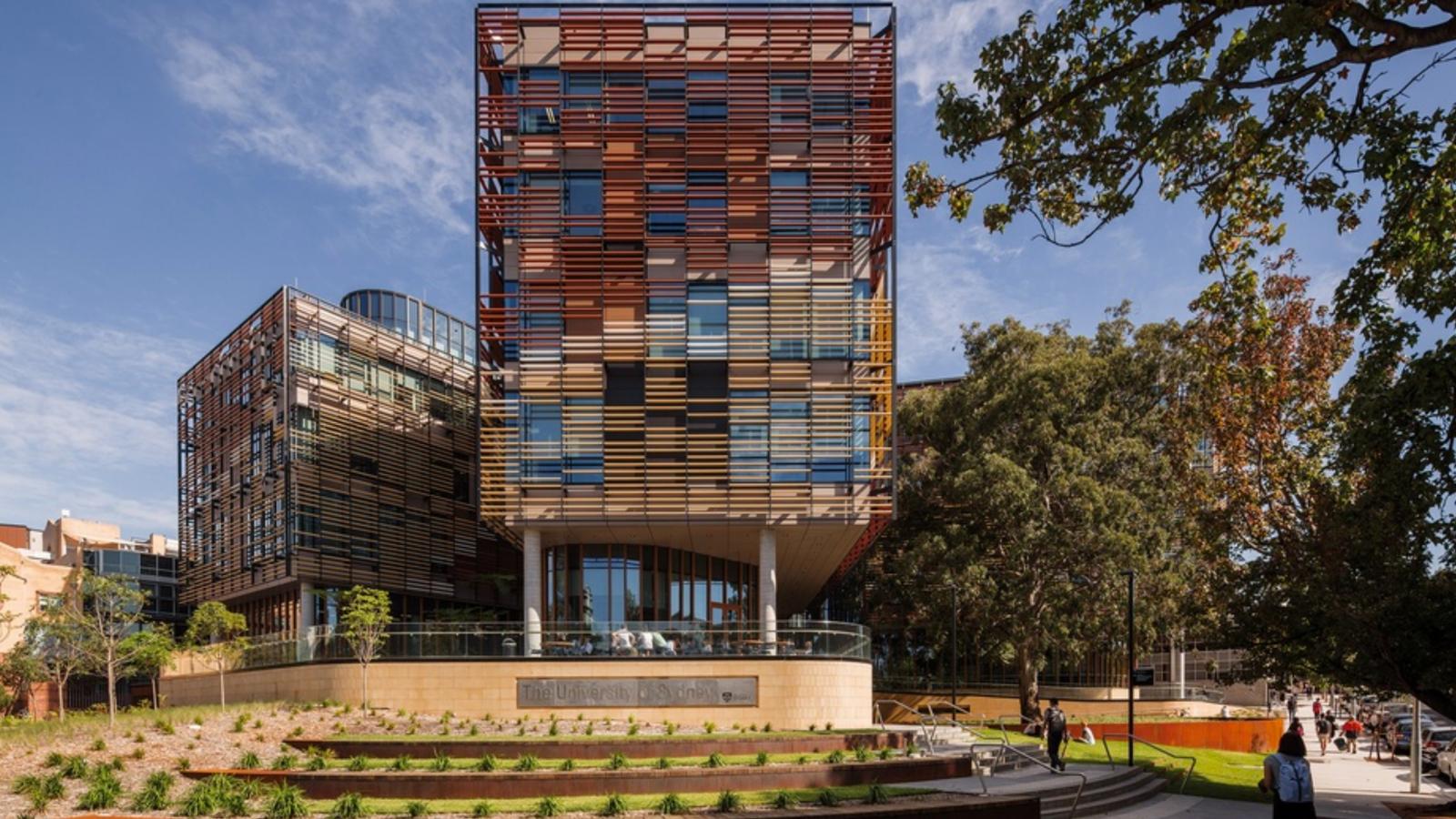 A modern building with a colorful, geometric facade made of horizontal and vertical panels, set on a lush, landscaped campus with trees and greenery. The structure features large windows and a sign that reads "University of Sydney Business School" at the entrance.