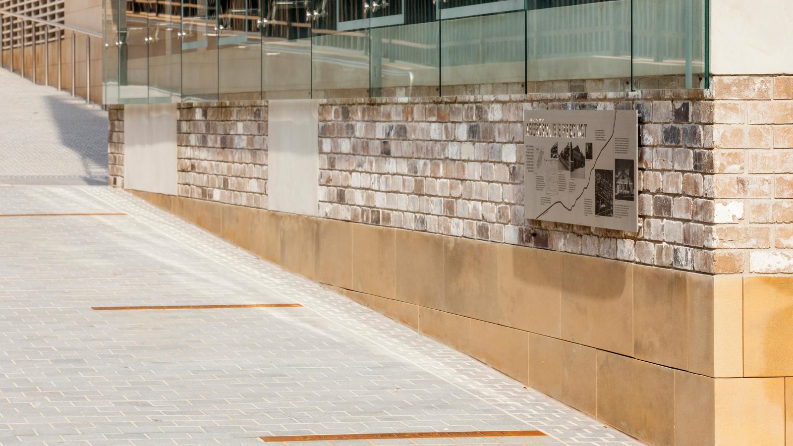 A modern brick and glass building belonging to the University of Sydney's Business School features a sidewalk and a metallic plaque mounted on the wall. The brickwork and large glass windows give the building a contemporary look while the sidewalk is paved with gray bricks.